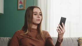 A young lady in a brown sweater and blue jeans sits on a sofa in front of a window and talks emotionally over a video call. Medium shot