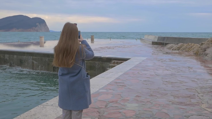 Young caucasian woman traveler takes a photo of waves crushing on the pier. Montenegro. Slow motion
