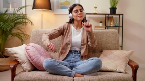 Young Caucasian Woman In Headphones Sitting In Couch In Bright Room At Home Listening Favorite Music And Dancing On Sofa, Raises Arms. Stay Home Quarantine Concept Of Relax And Meditation