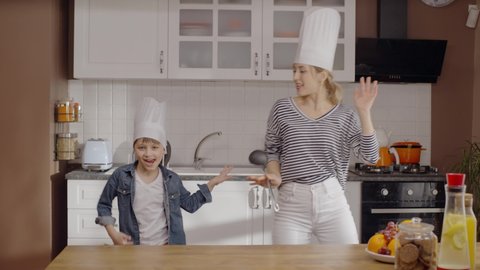 The young mother and her son dance wildly in the kitchen with the chef's hat on their heads, they sing and have a lot of fun together with the ladle in the mother's hand. Portrait of dancing family li