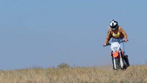 A girl on a sports motorcycle, quickly rides on dry grass against a blue sky, approaching the video camera. The girl participates in motorcycle competitions.