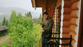 Serious man with a beard uses the Internet on a smartphone on the balcony of a hotel in the mountains on a rainy day. Rest in a country house.