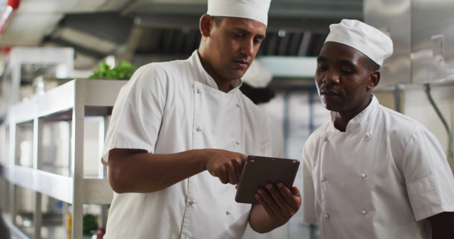 Two diverse male chefs talking and using tablet in restaurant kitchen. Working in a busy restaurant kitchen.