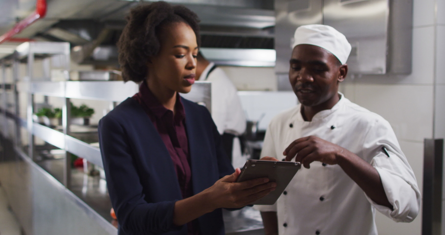 African american female manager using tablet and talking with chef. Working in a busy restaurant kitchen.