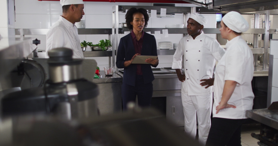 African american female manager using tablet and talking with chefs. working in a busy restaurant kitchen.