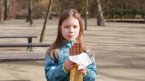 Little girl outdoors in the park eating a bar of chocolate biting and chewing - teeth health concept