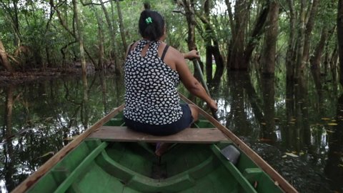 Amazonas, Brazil August 5, 2018: Native woman paddling a canoe in a river igarape between trees in the Amazon Rainforest. Concept of travel, environment, ecology, tourism, conservation, people.
