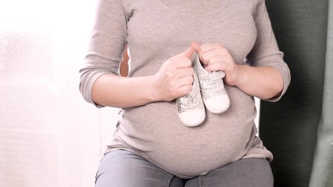 Young pregnant woman with small baby shoes sitting at home. Expectant mother is preparing for childbirth. Maternity and  pregnancy concept.   Pregnant woman stepping  baby booties over her big belly.