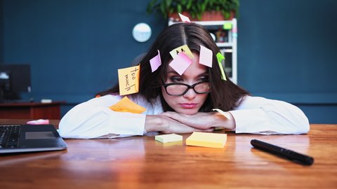 Crazy brown hair with lots post it stickers young freelance woman sit at the table in the office looking nowhere trying to work with blue color background. High quality 4k resolution footage. 