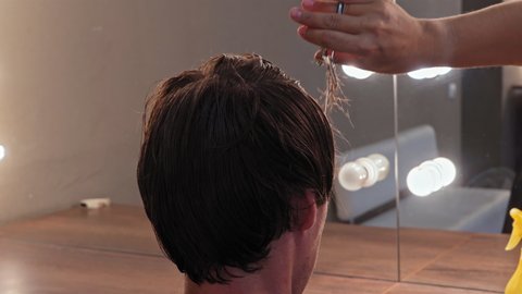 Close up of the back of the young man head with a hairdresser hand with scissors leveling the length of his hair against the background mirrors with lighting. High quality 4k resolution footage. 