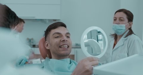 Arabian patient with open mouth sitting in the dentist chair, he is looking at the mirror and moving his jaw up and down after that dentist puts off from his mandible transparent plastic aligner.