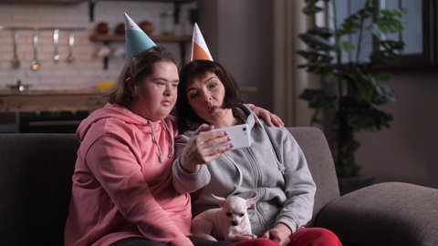 Joyful handicapped girl with down syndrome and her mother in cone hats singing happy birthday song while taking photo on cellphone. Positive disabled child with parent posing for selfie on sofa