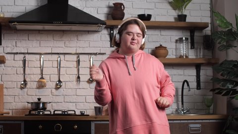 Close-up of disabled teen girl with down syndrome listening to music in headphones and dancing with her eyes closed in kitchen. Handicapped music lover in pink hoodie enjoying home leisure activity