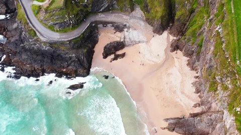 People on a stunning sand beach near dark rocky cliffs in Ireland. Aerial top down overhead view of strong ocean waves splashing ashore the hidden sand beach near Wild Atlantic Way
