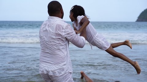 Father playing with daughter on the beach. they wear white suits. blue beach background with small waves. Dominican Republic. happy african american father and daughter