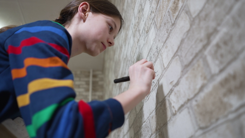 Smart girl wearing striped sweatshirt writing mathematic fractions on wallpaper with brick wall print. Low angle pupil doing homework in unusual way. Concept of education