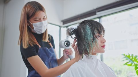 Professional stylist using hair dryer blow young woman's hair in salon after hair dyeing. hairdresser wearing mask to prevent from coronavirus infection during pandemic. Beauty salon business concept.