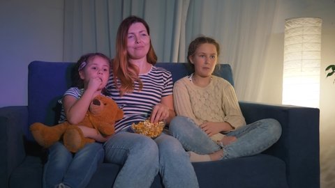 Happy family watching TV at home. Mom and kids are watching tv with popcorn. Happy family watching movie at home. Mom and children are eating popcorn on the couch. Happy family watching movie