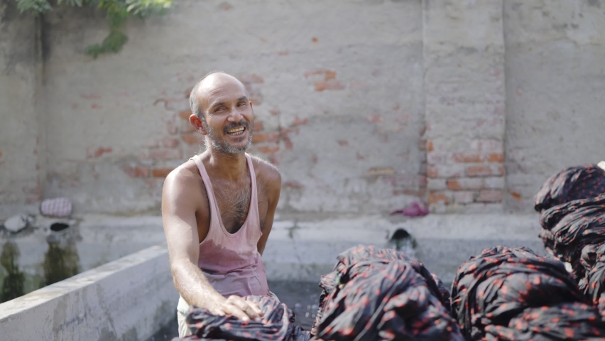 Shot of a happy looking middle aged Indian male daily wage laborer or washerman of local or domestic textile dyeing firm sitting next to wet dyed cotton sheets or garments and looking at the camera | Shutterstock HD Video #1068763463