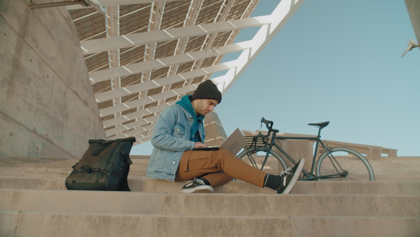 Modern millennial urban man sit on stairs of epic office building, work on laptop remotely from office. Freelance worker, young student work on remote project on the go. Commuter in city surroundings | Shutterstock HD Video #1068763541
