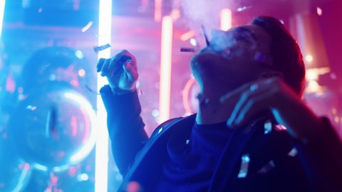 Closeup relaxed guy dancing under confetti at night club party. Portrait of hot male person moving with cigarette on neon lights background. Calm man blowing smoke in club.