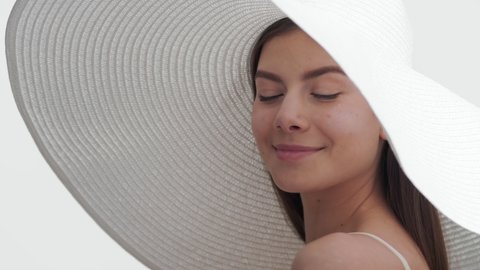 Young gorgeous European woman with long brown hair in a big white hat applies sunscreen on cheeks and smiles wide for the camera against white background | Sunscreen applying for face care commercial