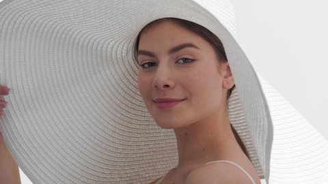 Young gorgeous dark-haired Caucasian woman in a big white hat enjoys the sun holding the brim of her hat, turns around and goes away against white background | UV protection concept
