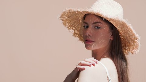 Young good-looking European woman with long dark hair in a straw hat and white bikini turns around touching her shoulder and smiling for the camera against beige background | Sun cream commercial