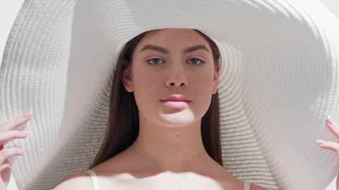 Young pretty brown-haired European woman in a big white hat holding the brim of it blows a kiss for the camera and smiles wide touching her face against white background | Face skin protection concept