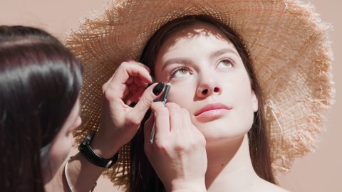 Brunette woman puts makeup on attractive European female model in a straw hat using eyeliner against beige background | Makeup putting on shot for skin care commercial