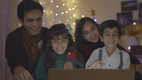 Shot of Indian Asian cheerful and close-knit family sitting on a couch talking on an online video call using laptop with great excitement at home well decorated on the occasion of Christmas eve