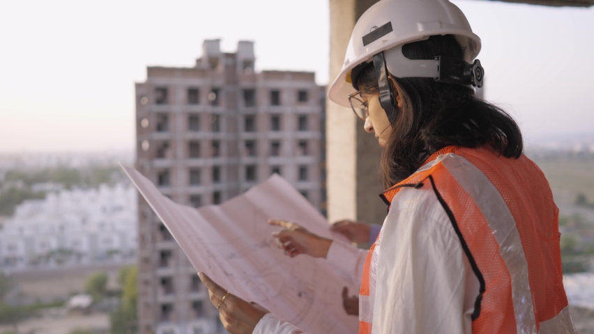 A young male and female Indian Asian civil engineers safety jackets and helmets standing on an under-construction building holding a blueprint of the structure and working together   Royalty-Free Stock Footage #1068774254