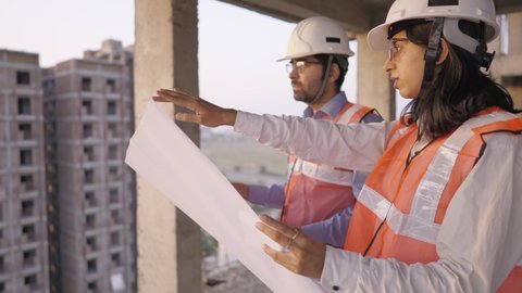 A young male and female Indian Asian civil engineers safety jackets and helmets standing on an under-construction building holding a blueprint of the structure and working together  
