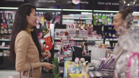 Medium side-view shot with slowmo of asian woman at cashier in makeup shop with Accessories, Deodorants and Shower Gels titles on stands, when shop assistant giving her bag with purchased products