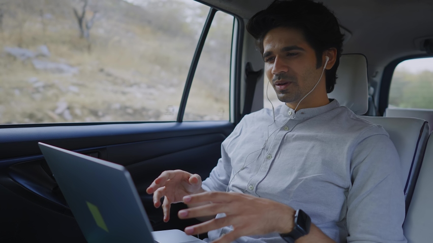 A shot of a young smiling and expressive Indian male entrepreneur having an online video call conversation on a  laptop computer while sitting in a rear seat of a moving car Royalty-Free Stock Footage #1068776423