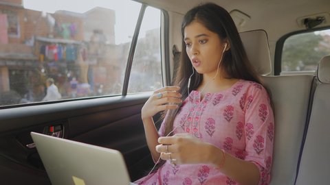 A young and beautiful corporate Indian female entrepreneur is busy having an online video conference meeting with office colleagues on a laptop sitting in a moving car through the busy city streets