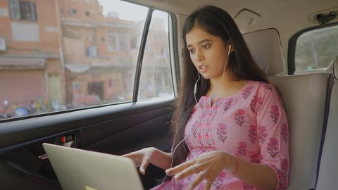 A young and beautiful corporate Indian female entrepreneur is busy having an online video conference meeting with office colleagues on a laptop sitting in a moving car through the busy city streets