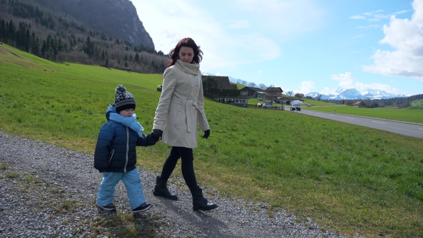 Mother with Child Walking Along Mountain Rural Road. Tourists Travel in Picturesque Swiss Alps. Scenic Landscape. Green Grass Pastures Meadows. Cottages in Village. Switzerland Europe