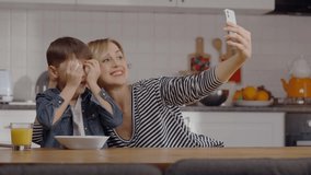 Young mother and her little son are taking selfie with a cell phone while cooking together in the kitchen. Mom and son having fun together.Slow motion video.