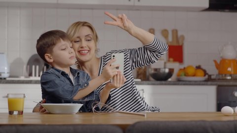 Young mother and her little son are taking a selfie with a cell phone while eating together in the kitchen. Mother makes victory sign (V). Mom and son having fun together. 
