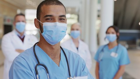 Confident male nurse in front of his medical team looking at camera wearing face mask during Covid19 pandemic. Smiling middle eastern surgeon standing in front of colleagues wearing protective mask.