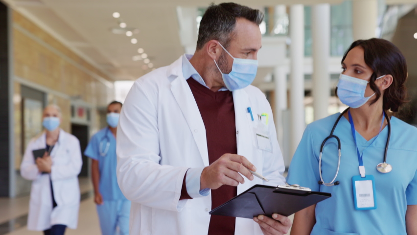 Mature doctor and nurse wearing safety mask for Covid-19 having a discussion in hospital hallway. Healthcare specialist showing medical report to nurse wearing face mask for protection against covid. | Shutterstock HD Video #1068792191
