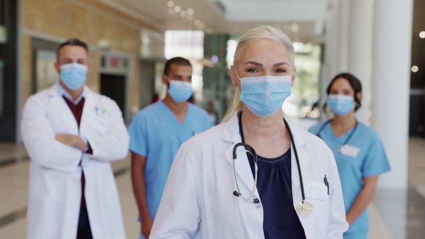 Portrait of smiling woman doctor standing in hospital with team in background wearing face mask during Covid-19 pandemic. Mature doctor with her medical staff looking at camera with surgical mask. | Shutterstock HD Video #1068792194
