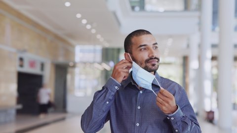 Indian businessman walking in a building while taking off protective face mask and looking at camera after immunization from Covid19. Middle eastern man removing mask from face after vaccine shot.