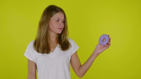 Woman in white T-shirt looks blue donuts in hand licks and bites lips on yellow background with copy space. 4K slow motion footage