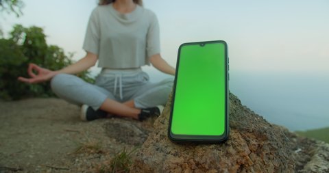 View of girl in yoga pose on mountain peak meditate with Green screen Mock up smartphone on foreground