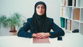 Muslim businesswoman smiling near paper folder and smartphone in office