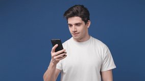 Cheerful young man 20s in white t-shirt posing isolated on blue color background in studio. People lifestyle concept. Talking on mobile cell phone making video call greeting with hand showing thumb up