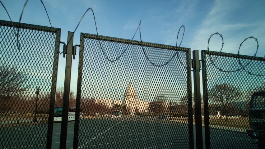 A time-lapse of the U.S. Capitol building at sunset, fortified behind a metal fence topped with razor wire to address security concerns following the January 6th 2021 insurrection. Camera dolly out. 