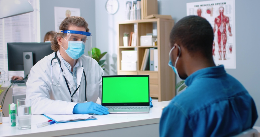 Rear of African American male patient sitting on consultation in clinic talking to Caucasian doctor in face shield and medical mask showing laptop with chroma key, monitor with green screen | Shutterstock HD Video #1068813329